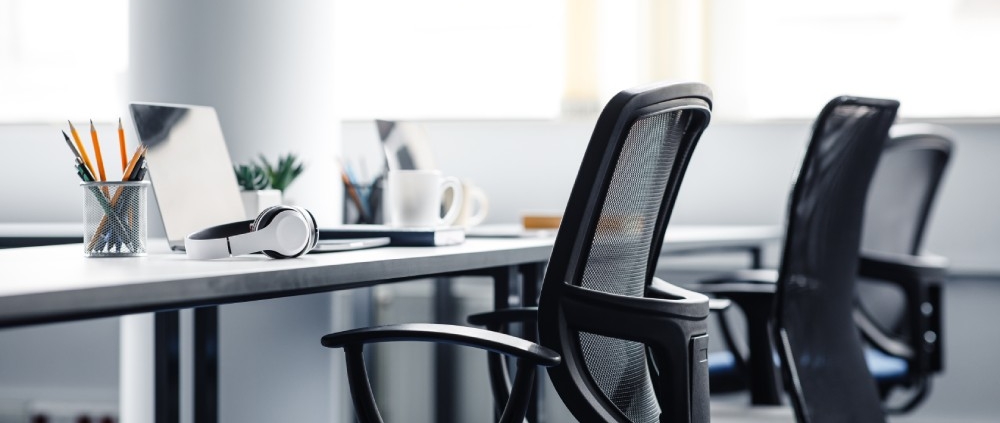 An office desk that features laptops, a cup of pencils, a coffee mug, headphones, and three ergonomic office chairs.