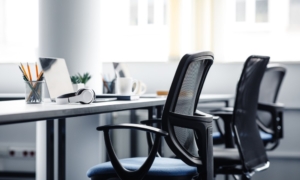 An office desk that features laptops, a cup of pencils, a coffee mug, headphones, and three ergonomic office chairs.