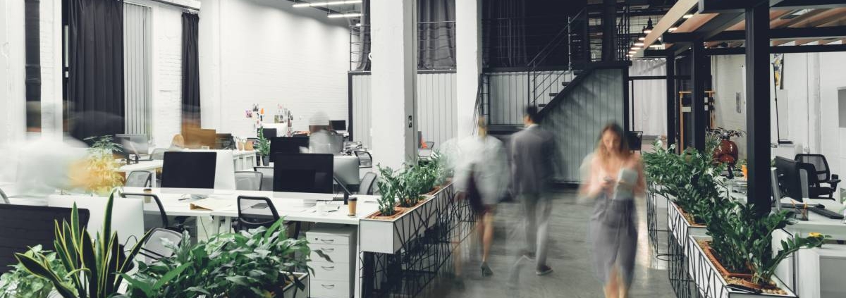 A modern-looking small office with an open layout, multiple workstations, and blurred business colleagues walking around.