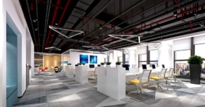 A modern office with open ceilings, carpeted floors, and rows of workstations with yellow chairs. Some windows show a city view.