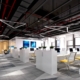 A modern office with open ceilings, carpeted floors, and rows of workstations with yellow chairs. Some windows show a city view.