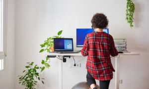 A woman in a red plaid shirt with her back to us, standing and working at a height-adjustable office desk, focused on her tasks.