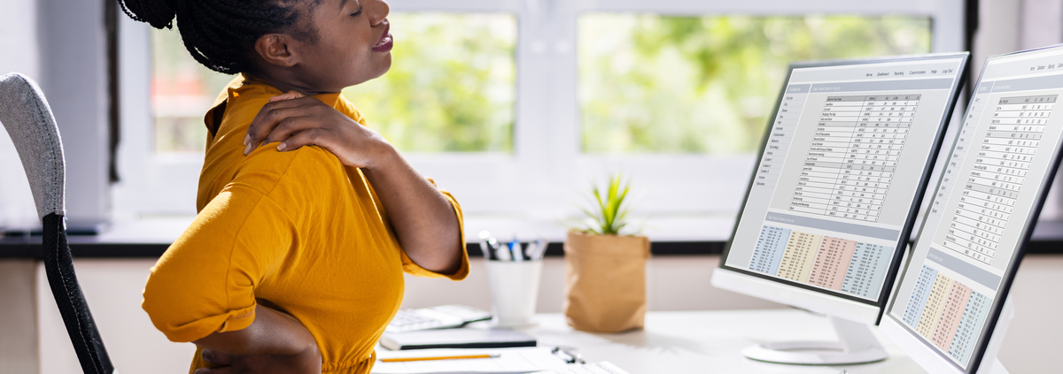 A woman sits at her desk, rubbing her neck and back with a pained expression, indicating discomfort and pain.