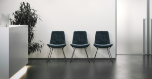 An office reception area with a white wall and dark floors. The reception area also has three chairs and a plant.