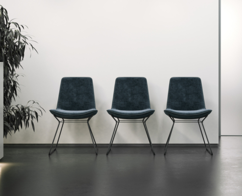 An office reception area with a white wall and dark floors. The reception area also has three chairs and a plant.