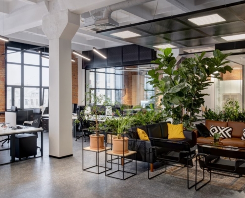 An office contains tables, chairs, plants, and a couch. The office has wide windows, and the layout is open.