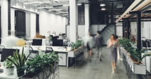 An open office with people walking around. There are multiple computer screens and desks with plants throughout.