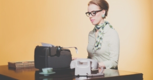 A woman sitting at a desk with a typewriter and a telephone. She's wearing glasses and a patterned scarf.