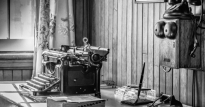 An office with a wooden desk, a typewriter, and a telephone. The desk also has a pen and a book on it.