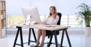 A woman sitting at her desk with her feet on a footrest. She has a monitor in front of her, and she's moving her mouse.