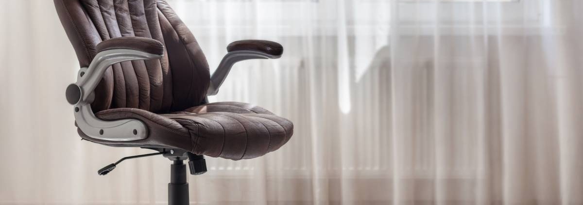 A brown leather office chair in a light-colored room next to a window. The sun is shining through the window.