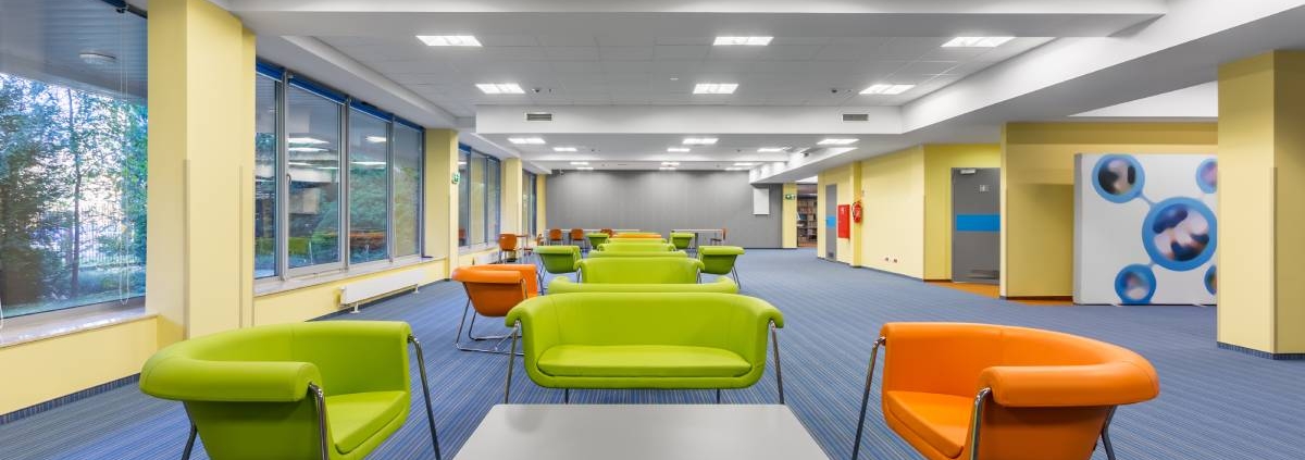 A large office space with blue carpet, yellow walls, green and orange chairs, green sofas, and gray tables.