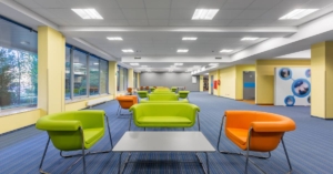 A large office space with blue carpet, yellow walls, green and orange chairs, green sofas, and gray tables.