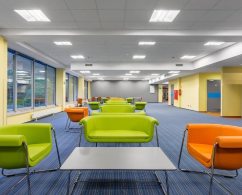 A large office space with blue carpet, yellow walls, green and orange chairs, green sofas, and gray tables.