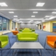 A large office space with blue carpet, yellow walls, green and orange chairs, green sofas, and gray tables.