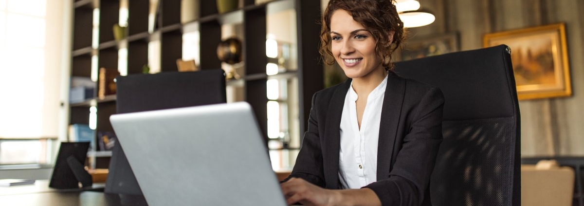 A woman working on a laptop while sitting at a desk in an office. She is looking at the laptop screen and smiling.