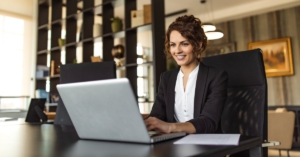 A woman working on a laptop while sitting at a desk in an office. She is looking at the laptop screen and smiling.