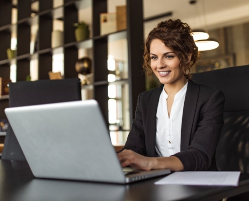 A woman working on a laptop while sitting at a desk in an office. She is looking at the laptop screen and smiling.