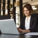 A woman working on a laptop while sitting at a desk in an office. She is looking at the laptop screen and smiling.