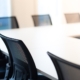 A row of office chairs around a table in a room. The office chairs are black, and there's a window in the background.