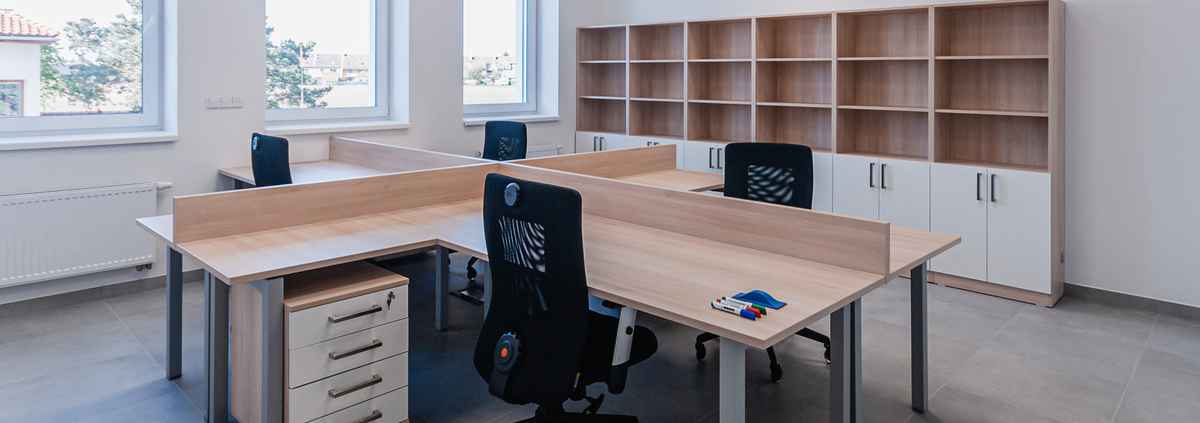 An office with a large shared desk and four office chairs. There are windows on one wall and a shelving unit along the other.