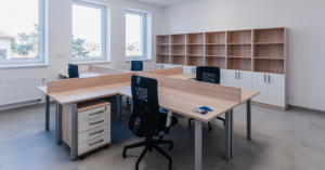 An office with a large shared desk and four office chairs. There are windows on one wall and a shelving unit along the other.