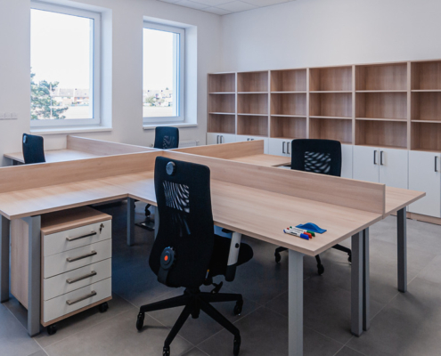 An office with a large shared desk and four office chairs. There are windows on one wall and a shelving unit along the other.