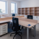 An office with a large shared desk and four office chairs. There are windows on one wall and a shelving unit along the other.