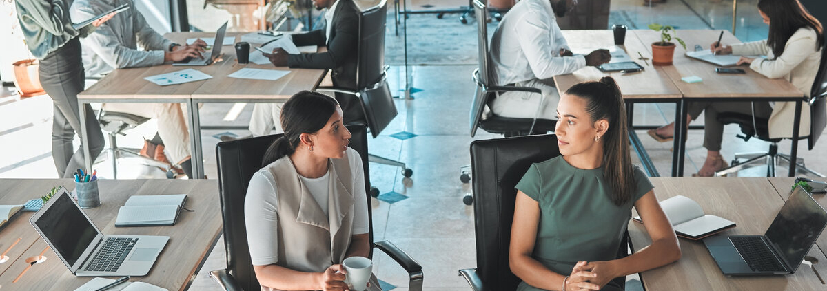 People sitting and standing in an open office. Two women face each other, having a pleasant-looking conversation.