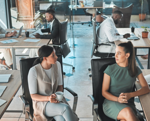 People sitting and standing in an open office. Two women face each other, having a pleasant-looking conversation.