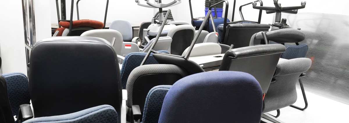 Multiple black, blue, and red office chairs are crowded in a brightly lit storage room with white walls.