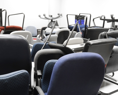 Multiple black, blue, and red office chairs are crowded in a brightly lit storage room with white walls.