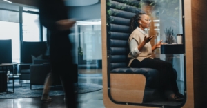 A young businesswoman smiling while working on a laptop inside of a glass and wooden privacy booth in a modern office.