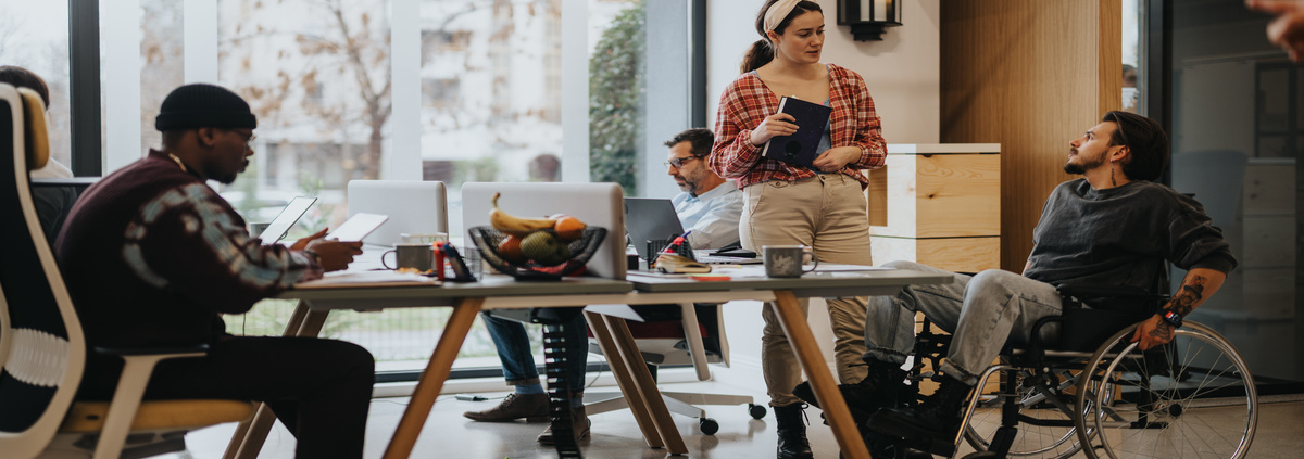 A diverse office team collaborates in an inclusive workspace, with one member in a wheelchair using a laptop.
