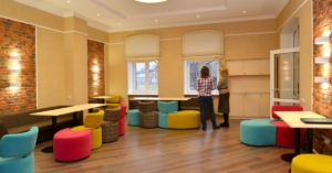 Two coworkers arrange colorful modular chairs at multiple desks in a brightly lit office workspace.