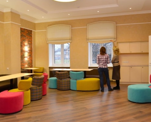 Two coworkers arrange colorful modular chairs at multiple desks in a brightly lit office workspace.