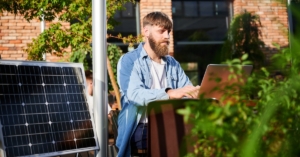 A man uses a laptop at an outdoor table with a large solar panel next to him in an eco-friendly workspace.
