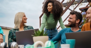 A group of smiling team members collaborate at an outdoor café terrace, using laptops in a shared lush green workspace.
