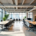 A modern open-plan office sits empty with shared desks, ergonomic chairs, and bright natural lighting.