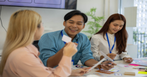 Three people sit at a table and collaborate on a project while reading information on two small white cards.