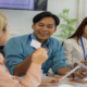 Three people sit at a table and collaborate on a project while reading information on two small white cards.