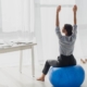 A businesswoman stretches her arms while sitting on a fitness ball in a bright, modern office workspace.
