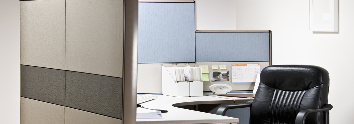 A black leather empty chair waits at tidy desk in cubicle that has blue shelves and white folder organizers.