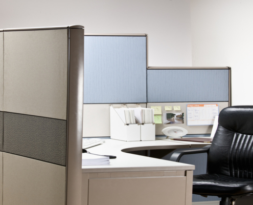 A black leather empty chair waits at tidy desk in cubicle that has blue shelves and white folder organizers.