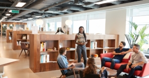 A group of casually dressed businessmen and businesswomen is holding a meeting in a modern, wooden-and-glass office.