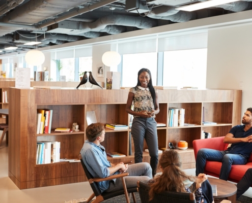 A group of casually dressed businessmen and businesswomen is holding a meeting in a modern, wooden-and-glass office.