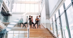 A group of young businesspeople standing on a staircase in a modern, glass building talking to each other.