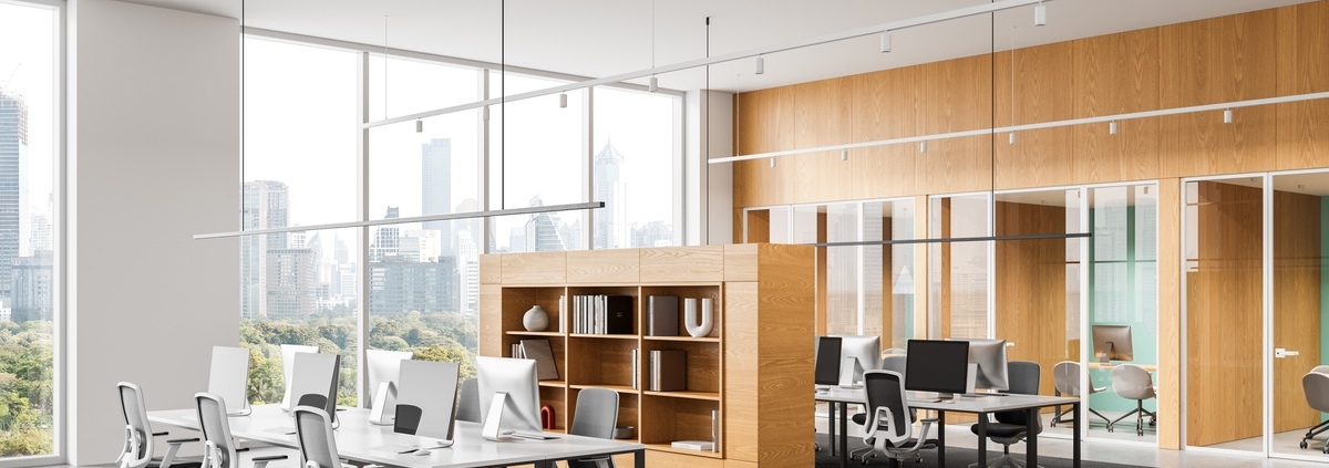 A modern open-plan office interior featuring shared desks, ergonomic chairs, wood partitions, and a cityscape view.