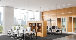A modern open-plan office interior featuring shared desks, ergonomic chairs, wood partitions, and a cityscape view.