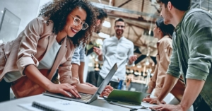 A group of young business people in an office, smiling, and gathered around their laptops while working.